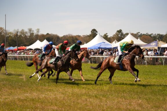 Horse racing event with jockeys in colorful attire riding thoroughbreds on a grassy track, spectators and tents in the background.