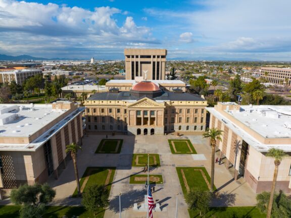 Aerial view of a historic building with a red dome and ornate facade, flanked by modern structures, green lawns, palm trees, with a background of a cityscape and mountains under a partly cloudy sky.