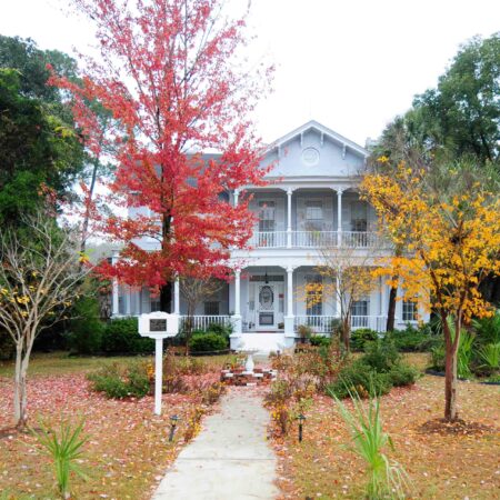 A two-story white house with a front porch, surrounded by autumn-colored trees and a leaf-covered lawn, with a path leading to the front entrance.