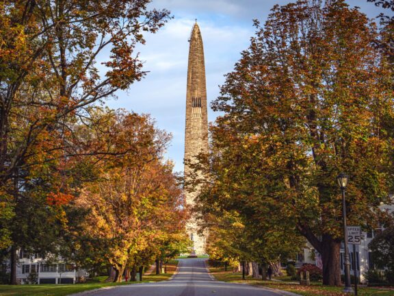 A picturesque street lined with autumnal trees leading towards a tall obelisk monument under a clear sky.