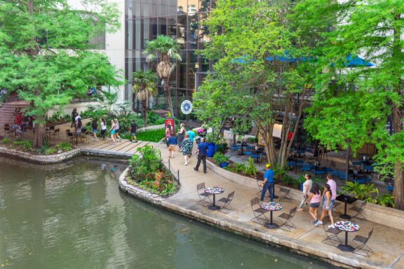 An outdoor urban scene with people walking along a riverside walkway, flanked by tables and chairs, green trees, and a glass-fronted building.