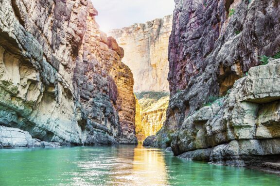 A tranquil river flows through a canyon with layered rock walls under a bright sky.