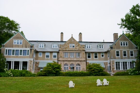 Large, traditional mansion with a stone facade and multiple gables, surrounded by a green lawn with two white Adirondack chairs in the foreground.