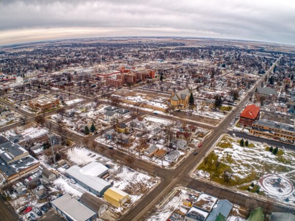 An aerial view of a snow-dotted town with a mix of residential and commercial buildings, streets forming a grid, and a flat horizon in the distance under a cloudy sky.