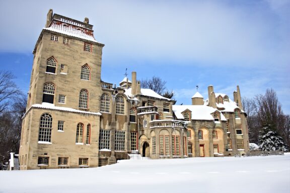 An ornate, historic mansion covered in snow, with multiple gables and a clear blue sky in the background.