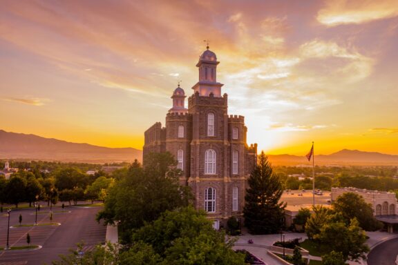 Sunset view of a historic multi-towered stone building with a United States flag in the foreground and mountains in the distance.