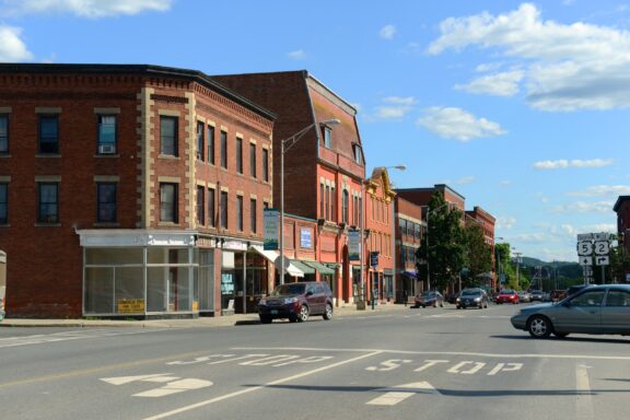 A sunny street scene with historic multi-story brick buildings on the left, a clear blue sky with scattered clouds above, and vehicles on the road featuring prominent 'STOP' road markings in the foreground.