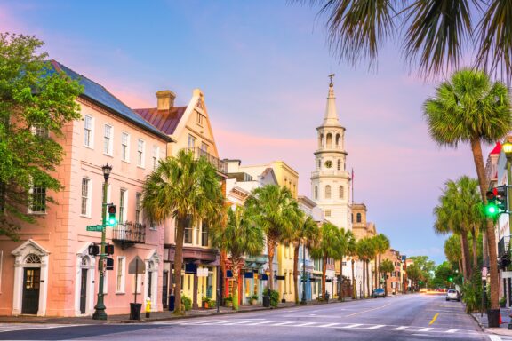 A picturesque view of a historic street lined with colorful buildings at dusk, featuring a prominent church steeple in the background and palm trees along the sidewalk.