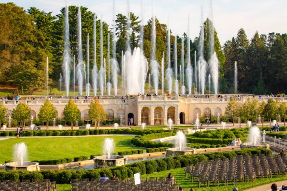 An ornate outdoor garden with a large fountain display, featuring multiple high jets of water, surrounded by neatly trimmed hedges, trees, and a classical architectural structure with visitors enjoying the views.