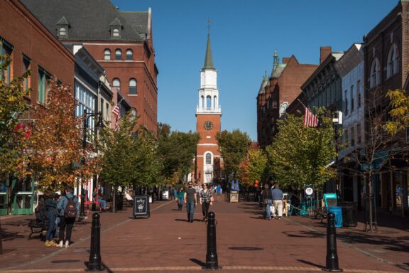 A bustling pedestrian street flanked by red brick and traditional buildings leading to a white-steepled church under a clear blue sky.