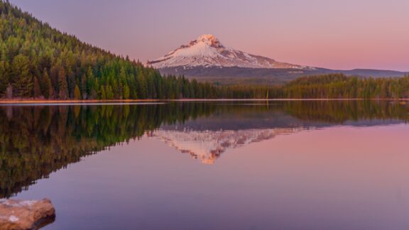 A serene view of a snow-capped mountain peak during sunset with colorful skies reflected in the still waters of a forest-lined lake.