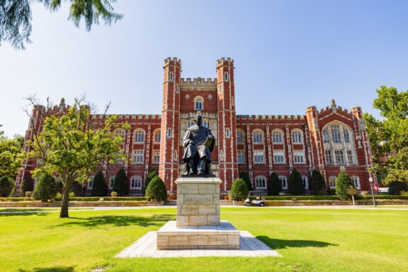 A view of a stone statue and the brick façade of the University of Oklahoma on a sunny day.