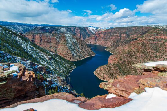 Scenic view of a snow-dusted canyon with a meandering river, red rock formations, and a partly cloudy sky.