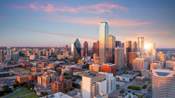 A panoramic cityscape at sunset with a mix of modern high-rise buildings and historical architecture, prominent skyscrapers reflecting the sun's rays, under a sky with soft pink clouds.