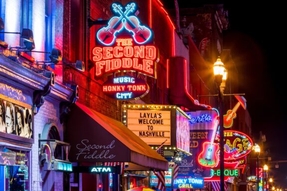 A vibrant nighttime street scene featuring neon signs of bars and restaurants with music and honky-tonk themes, including guitars and welcoming messages, indicative of a lively downtown Nashville.