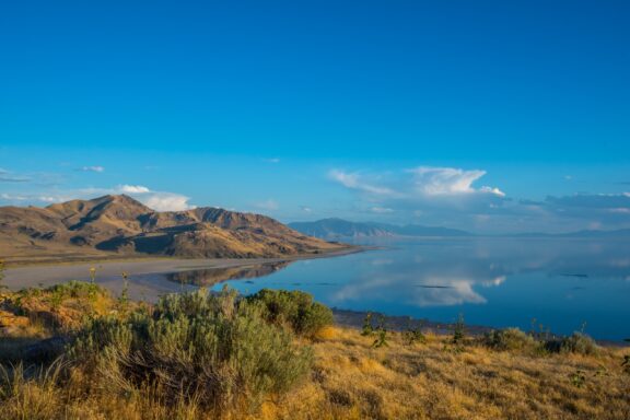 A serene landscape with a calm lake reflecting the blue sky and clouds, surrounded by mountains and vegetation in the foreground.