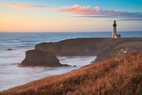 Sunset at Yaquina Head Lighthouse in Oregon with Coastal Cliffs and Ocean Waves A serene landscape featuring a lighthouse on a grassy cliff overlooking the ocean at sunset, with wispy clouds in the sky above and gentle waves below.