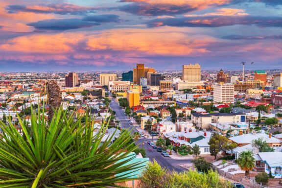 A vibrant sunset over a cityscape with colorful clouds, high-rise buildings in the center, residential areas in the foreground, and a green yucca plant on the left.