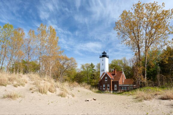 A white lighthouse with an attached red-roofed building surrounded by trees with early autumn foliage against a blue sky with wispy clouds. The foreground shows a sandy beach with grass tufts.