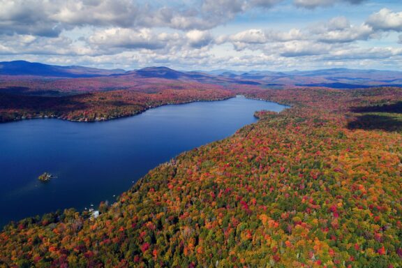 Aerial view of a lake surrounded by a colorful autumn foliage with mountains in the background under a partly cloudy sky.