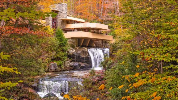 A modern house with horizontal lines built over a waterfall, surrounded by a forest with autumn-colored leaves.