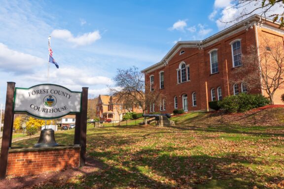 A sunny view of the Forest County Courthouse with a sign and bell in the foreground, the American flag, and a historic red brick building amid autumn foliage.