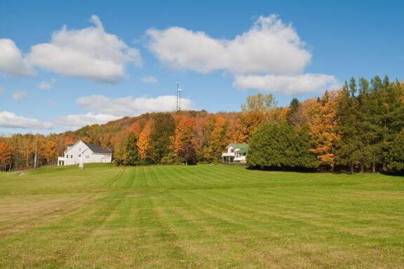 A picturesque rural landscape featuring a lush green field in the foreground with two white houses, surrounded by trees with autumn foliage, under a blue sky with scattered clouds.