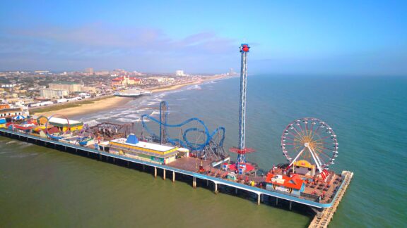 Aerial view of a vibrant amusement park on a pier with a roller coaster, Ferris wheel, and other attractions, extending into the sea with a coastal cityscape in the background.