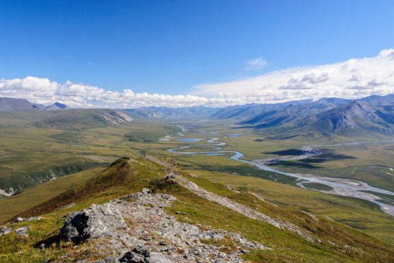 A panoramic view of a vast valley with a meandering river, flanked by rolling green hills and distant mountains under a blue sky with scattered clouds.