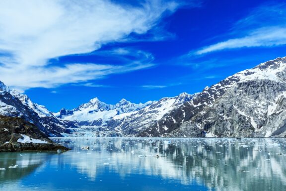 A serene glacial landscape featuring snow-capped mountains, a vibrant blue sky with wispy clouds, and a calm, reflective icy water body with floating ice chunks.