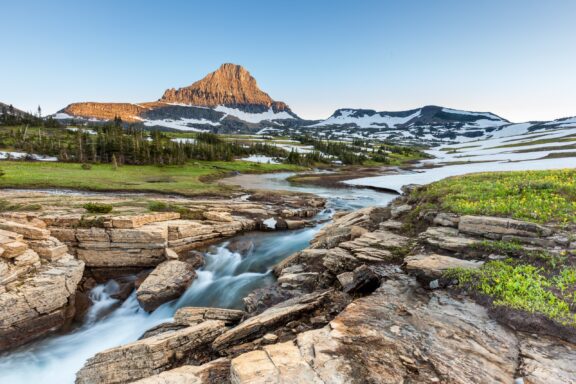 A scenic view of a rugged mountain peak during golden hour, with a river cascading over layered rocks in the foreground, patches of snow and vibrant wildflowers dotting the alpine landscape in one of the largest national parks in the US.