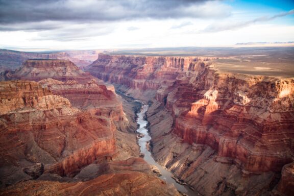 A panoramic view of the Grand Canyon with the Colorado River winding through the reddish-brown stratified rocks under a cloudy sky.