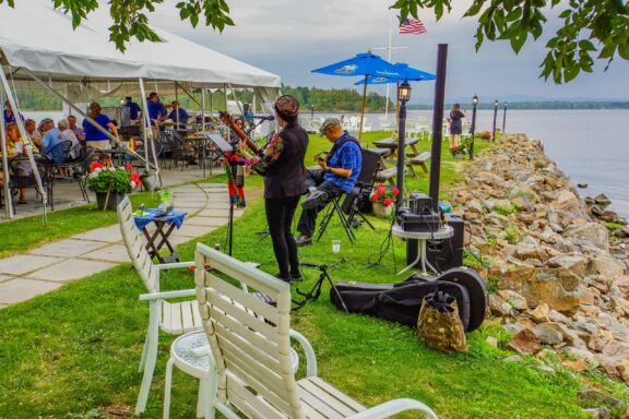 Musicians perform outdoors near a lakeside, with an audience in a tented area behind them, white chairs in the foreground, and rocky shoreline leading to a calm lake under a cloudy sky.