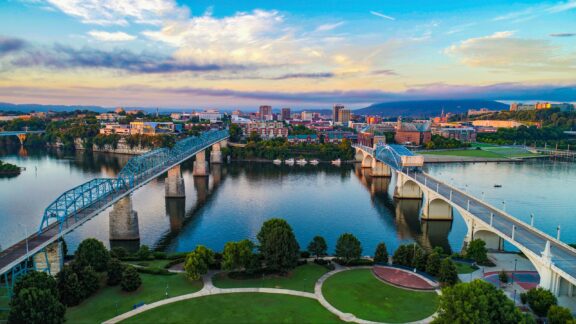 Aerial view of a cityscape at sunset with two bridges spanning a calm river, parks in the foreground, and a colorful sky in the background.