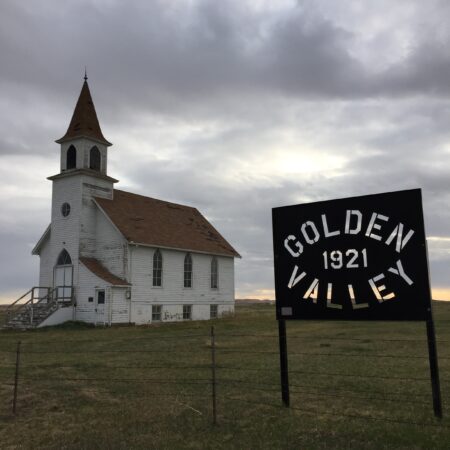 An old white church with a steeple stands under a cloudy sky beside a sign that reads "GOLDEN VALLEY 1921" on an open grassy field with a barbed wire fence in the foreground.