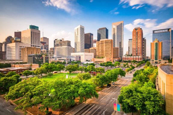 A scenic cityscape with modern high-rise buildings, lush green trees in the foreground, and a clear blue sky with fluffy clouds.