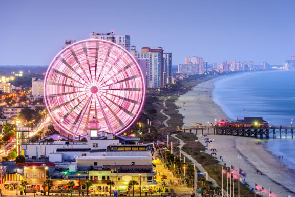 A dusk view of a lit Ferris wheel by the coast, with a beachfront, pier, and city buildings extending into the distance.