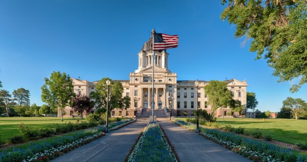 A stately capitol building with a large American flag flying atop under clear blue skies, surrounded by manicured lawns and colorful flowerbeds along a central walkway.