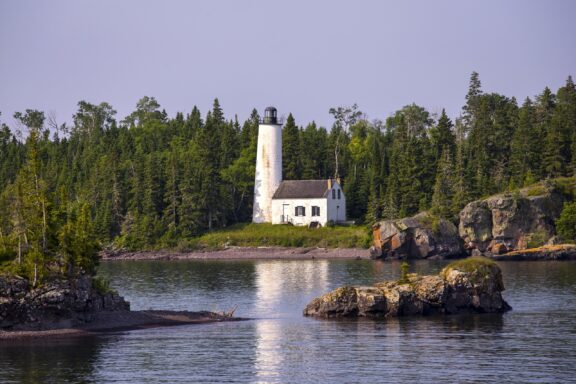 A white lighthouse with an attached keeper's house, surrounded by dense green coniferous trees and rocky outcrops.