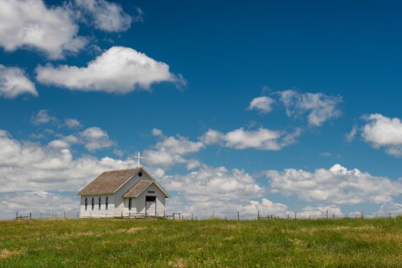 A small white church with a cross on top sits in an open grassy field under a blue sky with scattered clouds.