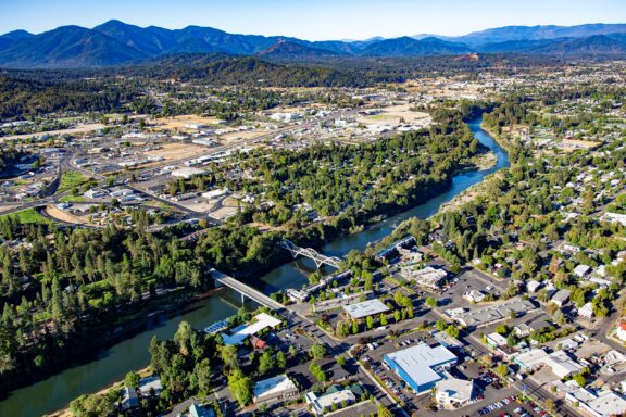 Aerial view of Grants Pass and the river running through it, with bridges connecting tree-lined streets amidst residential and industrial areas with mountains in the background.