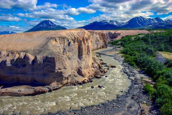 A winding river cuts through a colorful landscape with a steep, eroded canyon on one side and lush greenery on the other, set against a backdrop of majestic snow-capped mountains under a cloud-dotted blue sky.