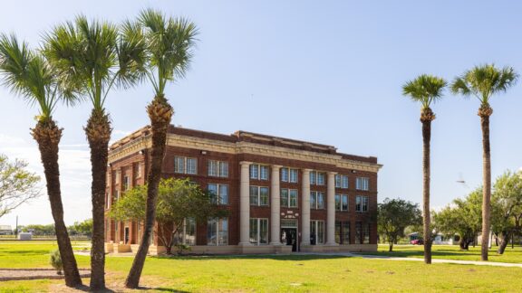 A classic red brick building with white trim and columns, framed by tall palm trees under a clear blue sky.