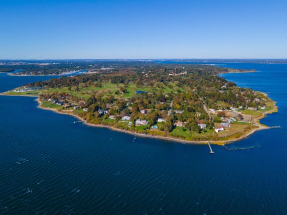 Aerial view of a coastal peninsula with residential housing, green spaces, and a dock extending into blue waters, under a clear sky.