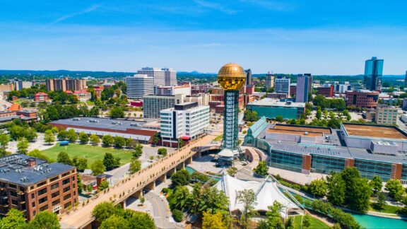 Aerial view of a sunny urban landscape featuring a unique golden sphere atop a tower, with a mix of modern and traditional buildings, green spaces, and a river running through the city.