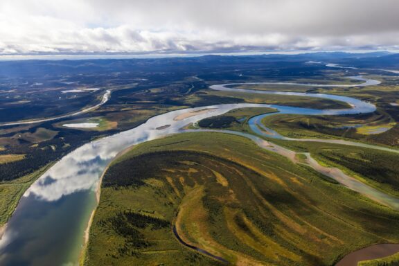 An aerial view of a meandering river flowing through a vast landscape with mixed forest and open areas, under partly cloudy skies.