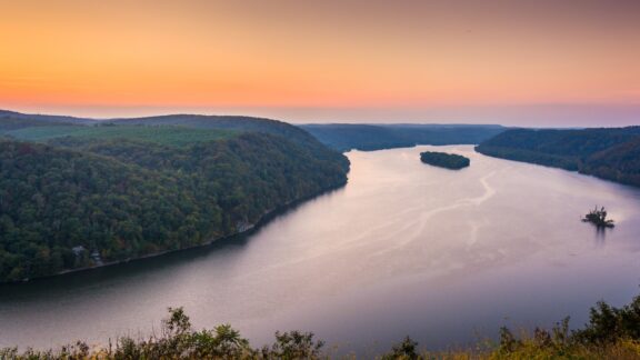 A serene sunset over a winding river flanked by forested hills, with a small island visible in the distance.