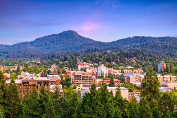 A scenic view of the Eugene cityscape with modern buildings surrounded by lush green trees at the base of a hill covered in dense forest.