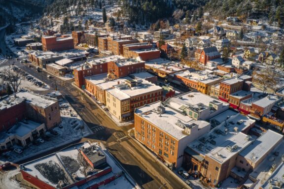 An aerial view of a snow-covered small town with traditional buildings, roads, and scattered trees on a sunny day.