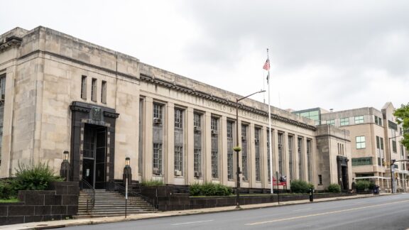 Exterior view of a historic United States Post Office building with neoclassical architectural elements and the American flag flying overhead, located on a city street corner.